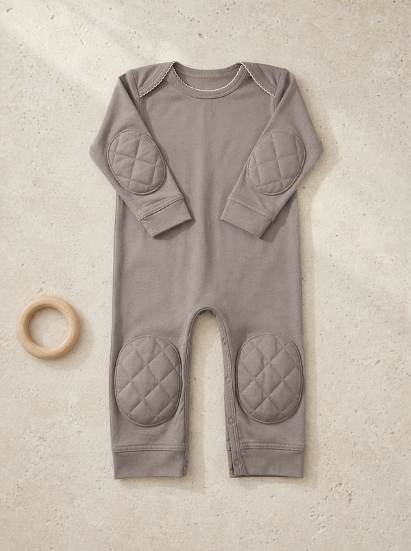 Grey padded onesie flat lay on warm stucco texture with wooden teething ring - front view showing quilted knee and elbow pads