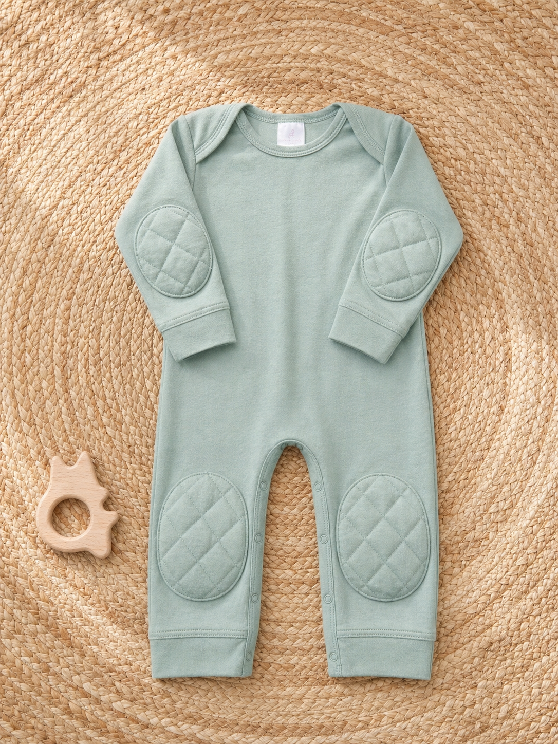 ComfyCrawlers Green Tea padded onesie laid flat on a woven seagrass placemat with wooden teething ring - showing full garment proportions